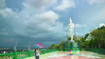 Thailand Chonburi 2023 september 9 Tourist holding a rainbow umbrella at Wat kho pra kru temple white buddha stand and dark gray cloud in rainy video