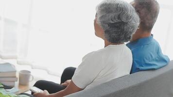 content Sénior couple en utilisant éloigné contrôle à changement canaliser. les personnes âgées regarder la télé pour divertissement tandis que homme en utilisant éloigné contrôle. épouse et vieux homme séance sur canapé dans vivant pièce avec télévision montrer. video