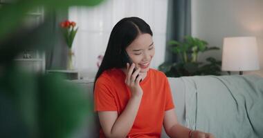 Footage of Young Asian woman talking with friend on mobile and reading a book while sitting on the sofa in the living room. Wellness at home, relaxing and lifestyle concepts. video