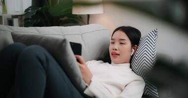 Footage of Happy young Asian woman listening to music on a smartphone while lying on the sofa in the living room. Wellness at home, relaxing and lifestyle concepts. video