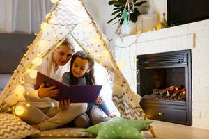 Mother and daughter are sitting in a teepee tent, reading stories with the flashlight. Happy family. photo