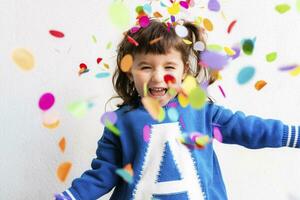 contento pequeño niña soplo el papel picado a un fiesta en frente de un blanco pared foto