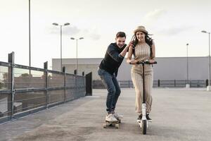 Young man and woman riding on longboard and electric scooter on parking deck photo