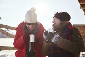 Happy mature couple with hot drinks outdoors at mountain hut in winter photo