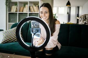 Smiling beautiful female influencer sitting on sofa while vlogging through smart phone at home photo