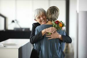 contento mayor mujer de negocios con manojo de flores abrazando colega en oficina foto