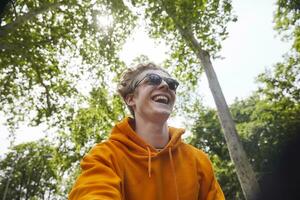 retrato de riendo joven hombre vistiendo Gafas de sol y naranja capucha camisa en naturaleza foto