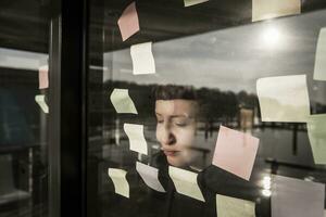 Tired woman leaning against window pane with sticky notes photo