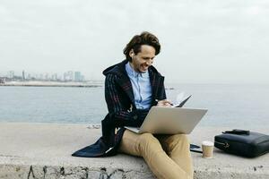 Spain, Barcelona, smiling man sitting at the sea working with laptop and notebook photo