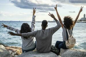 Three friends sitting on the beach, lookin at the sea, cheering, rear view photo