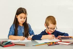 dos sonriente pequeño niños a el mesa dibujar con lápices de color, aislado en blanco foto