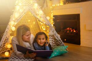 Mother and daughter are sitting in a teepee tent, reading stories with the flashlight. Happy family. photo