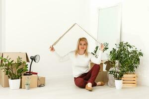 Happy young woman sitting on floor near moving boxes. Young woman moving to new home. Woman looking at camera and smiling photo