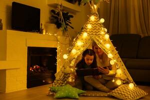 Mother and daughter are sitting in a teepee tent, reading stories with the flashlight. Happy family. photo