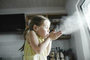 pequeño niña soplo harina en el aire en el cocina foto