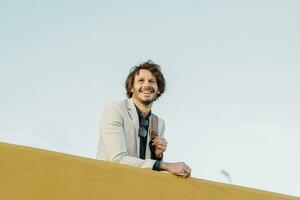 Portrait of laughing businessman with backpack leaning on wall looking at distance photo