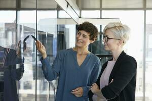 Two smiling businesswomen looking at chart on glass pane in office photo