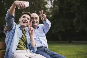 Senior man and grandson sitting together on a park bench taking selfie with smartphone photo