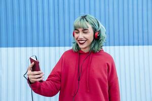 Portrait of laughing young woman with blue dyed hair with headphones taking selfie with mobile phone photo
