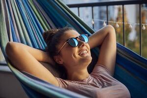 Portrait of happy young woman with sunglasses lying on hammock on balcony photo