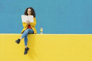 Young woman sitting on yellow wall reading a book photo