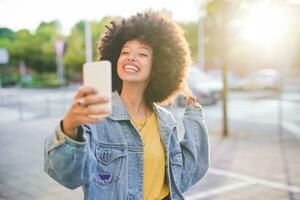 contento joven mujer con afro peinado tomando un selfie en el ciudad foto