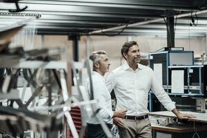 Mature colleague discussing with smiling manager while standing by manufacturing machinery in factory photo