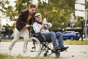 Smiling young man pushing happy senior man with smartphone in wheelchair photo