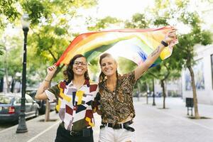 Happy lesbian couple waving rainbow flag while standing on footpath in city photo