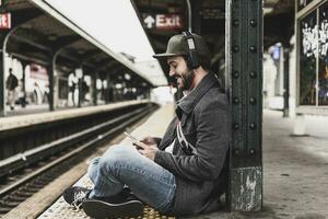 Young man waiting for metro at train station platform, using smart phone photo