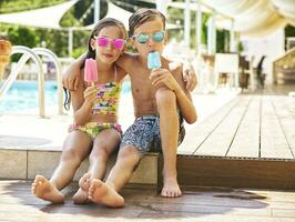 Portrait of little girl and boy with popsicles wearing mirrored sunglasses in front of swimming pool photo