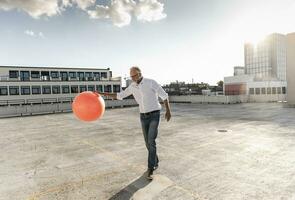 Mature man playing with orange fitness ball on rooftop of a high-rise building photo