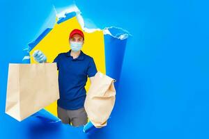 Delivery man in uniform hold craft paper packet with food isolated on blue background. Male employee in cap t-shirt working as courier. Service concept. Mock up copy space photo