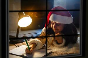 Little girl in Santa hat writes letter to Santa Claus near christmas tree and clock through the window photo