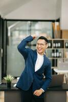 Young attractive Asian man smiling thinking planning writing in notebook, tablet and laptop working from home, looking at camera at office photo