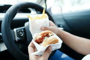Asian woman driver hold and eat hamburger and french fries in car, dangerous and risk an accident. photo