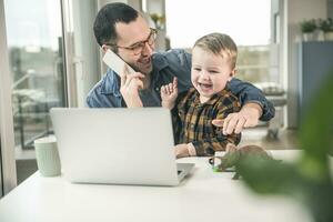 Happy father on cell phone in home office with son sitting on his lap photo