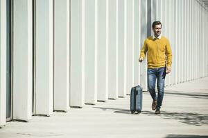 Young man with rolling suitcase and earphones in the city on the go photo