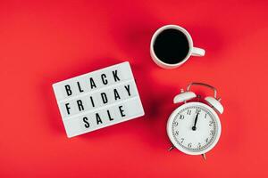 Black friday sale word on lightbox, cup of coffee, alarm on red background table. Flat lay photo