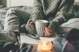 Female hands holding cozy mug with cacao or chocolate. Woman in gray knitted sweater sitting on bed. photo
