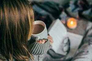 Young girl drinking winter beverage hot cacao, sitting on cozy bed, reading book. Cozy warm image photo