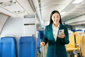 Portrait of  successful Asian businesswoman or entrepreneur in a formal suit on an airplane sitting in business class using a phone, computer laptop. Travel in style, work with grace. photo