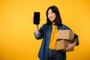 Portrait asian young woman wearing yellow t-shirt and denim shirt holding mobile phone and parcel box isolated on yellow studio background, Delivery courier and shipping service concept. photo