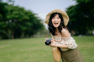 retrato de asiático joven mujer viajero con Costura sombrero y cesta y un cámara en verde público parque naturaleza antecedentes. viaje viaje estilo de vida, mundo viaje explorador o Asia verano turismo concepto. foto