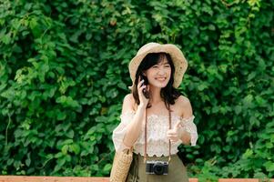 Portrait of asian young woman traveler with weaving hat, basket, mobile phone and camera on green public park background. Journey trip lifestyle, world travel explorer or Asia summer tourism concept. photo