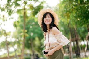 Portrait of asian young woman traveler with weaving hat and basket and a camera on green public park nature background. Journey trip lifestyle, world travel explorer or Asia summer tourism concept. photo