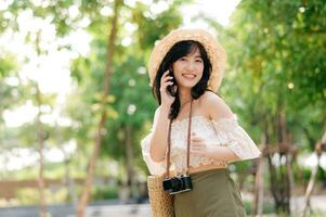 Portrait of asian young woman traveler with weaving hat, basket, mobile phone and camera on green public park background. Journey trip lifestyle, world travel explorer or Asia summer tourism concept. photo