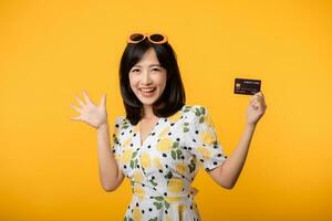 retrato joven asiático mujer contento sonriente en primavera vestir demostración el plastico crédito tarjeta aislado en amarillo antecedentes. pagar, dinero y compra compras pago concepto. foto