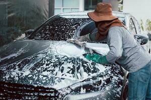 Woman worker washing car at car station outdoors with foam and yellow sponge. photo