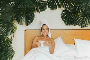 Woman lying in bed with green plant holding coffee mug with a towel on her head. photo